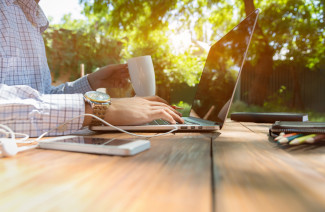 男人坐在自然乡村风格的木桌，周围有电子设备，在笔记本电脑上工作，喝咖啡，阳光和绿色露台的背景
